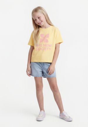 Mädchen mit langen blonden Haaren, das ein gelbes T-Shirt mit Grafik, hellblaue Jeansshorts und weiße Turnschuhe trägt, stehend und lächelnd.