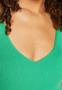 Tessuto verde a coste con scolatura a V profonda. La trama appare morbida e leggermente elastica, adatta a un indosso aderente. Presenta capelli ricci da un lato.