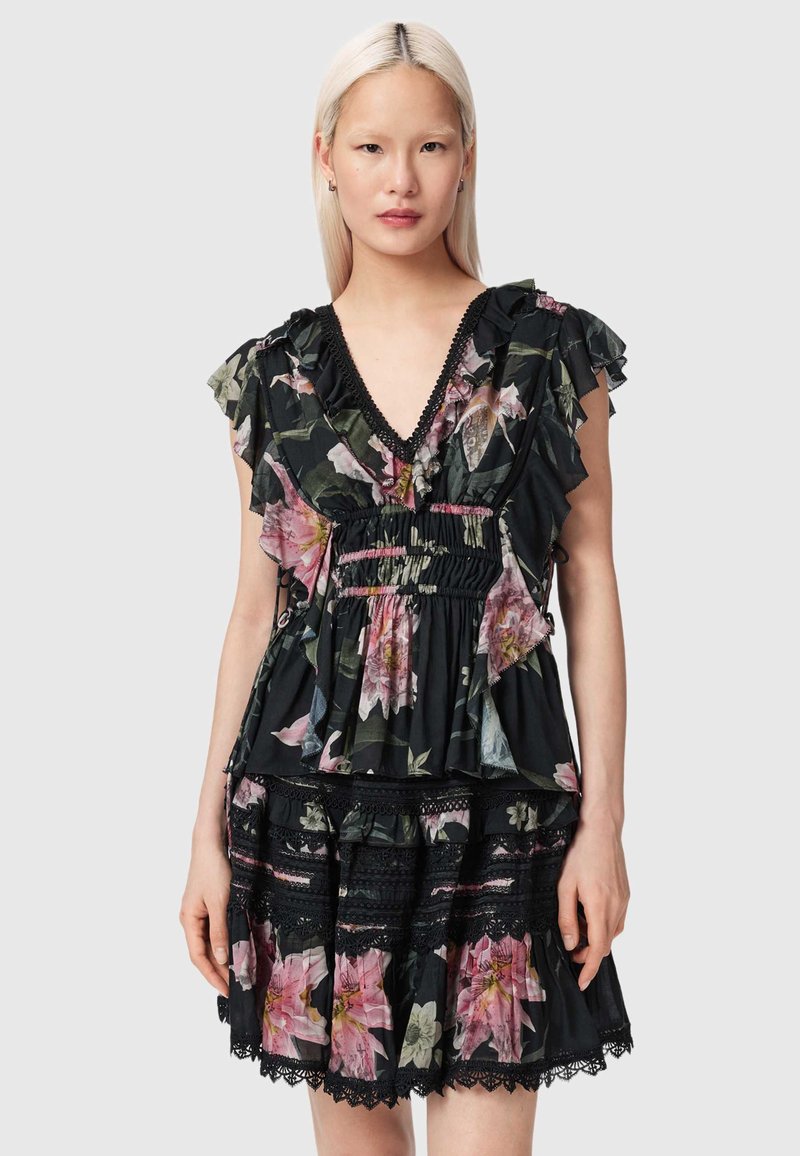 Femme aux longs cheveux blonds portant une robe noire à fleurs avec des volants, des détails en dentelle et un décolleté en V, debout contre un fond clair uni.