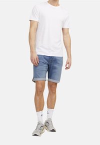 Valkoinen puuvillapaita, siniset farkkushortsit, joissa on käännetyt lahkeet, valkoiset urheilusukat ja harmaat tennarit, seisoo yksinkertaisella taustalla.