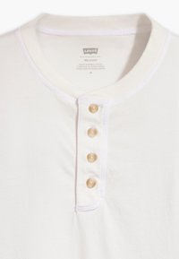 Maglietta henley bianca a vestibilità rilassata realizzata in tessuto morbido, con colletto rotondo e cinque bottoni color crema sul davanti.