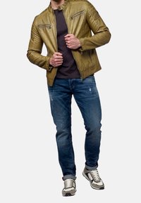 Giacca in pelle verde oliva con colletto stand, dotata di tasche con zip e dettagli testurizzati, abbinata a jeans blu strappati e scarpe da ginnastica multicolori.