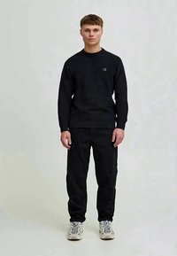 Tânăr stând îmbrăcat cu un pulover cu dungi negre, pantaloni negri și adidași albi, pe un fundal simplu de gri deschis.