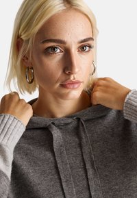 Šedá mikina s kapucí a pružnými manžetami. Ruka modelu tahá za kapuci, přičemž ukazuje měkkou strukturu a pohodlný střih na jednolitém pozadí.