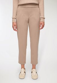 Beżowe spodnie taperowane o miękkiej teksturze, z przodu posiadające zakładki. W połączeniu z jasnobeżowymi sneakersami, oferującymi swobodny, nowoczesny wygląd.