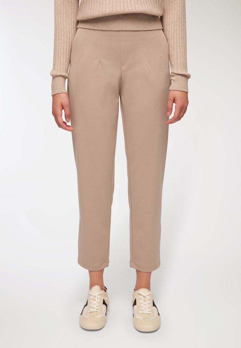 Beżowe spodnie taperowane o miękkiej teksturze, z przodu posiadające zakładki. W połączeniu z jasnobeżowymi sneakersami, oferującymi swobodny, nowoczesny wygląd.