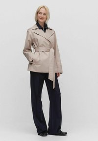 Beżowy trench z paskiem w talii, szerokimi klapami i długimi rękawami, zestawiony z granatowymi spodniami w prążki i czarnymi butami. Gładka tekstura.