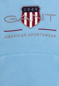 Felpa azzurra chiara con un grande logo ricamato che presenta un design a scudo, strisce rosse e bianche, e il testo "GANT American Sportswear."