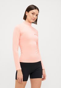 Maglia a maniche lunghe da surf di colore rosa chiaro, con colletto alto e dettaglio del logo. Il materiale è liscio ed elastico. Abbinata a pantaloni corti neri.