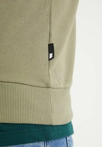 Gros plan d’un sweat-shirt beige à ourlet côtelé sur une chemise verte et un jean bleu, montrant une petite étiquette noire avec un carré blanc.