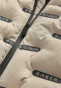 Beige gepolsterter Stoff mit schwarzem Reißverschluss und wiederholt aufgedruckten "TED BAKER"-Logos in schwarzen ovalen Formen.