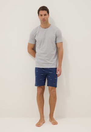 Man wearing a light gray short-sleeve shirt and navy blue shorts with small white sailboat patterns, standing barefoot against a plain wall.