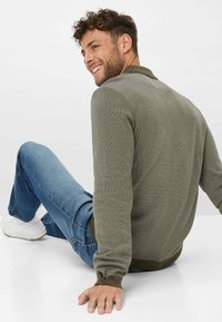 Khaki-grüner Rippstrickpullover mit Kragen, langen Ärmeln und tailliertem Saum, kombiniert mit blauen Jeans und weißen Sneakern. Sitzende Position.