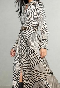 Abito camicia in beige con strisce nere diagonali a contrasto e motivi geometrici, caratterizzato da una vita con cintura e maniche lunghe.
