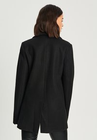 Blazer noir ajusté avec une coupe cintrée, une fente unique à l'arrière et des détails de boutons sur les manches. Fabriqué en tissu en laine mélangée texturée.