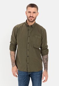 Olijfgroene button-up shirt met een enkele borstzak, opgerolde mouwen en donkere knopen. Draagt het met blauwe spijkerbroeken en zichtbare tatoeages.