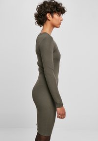 Robe longue à manches longues côtelées en vert olive, coupe près du corps avec un col rond et une fente sur le côté, mettant en valeur un tissu texturé et une silhouette élégante.