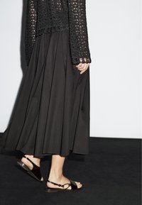 Top nero in pizzo a maniche lunghe abbinato a una gonna scura plissettata e sandali marroni, che mettono in risalto il tessuto strutturato e le morbide pieghe.