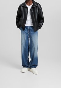 Giacca nera in similpelle, t-shirt bianca, jeans a gamba larga blu sbiaditi e sneakers bianche. La giacca presenta un'apertura frontale a pressione e polsini elasticizzati.