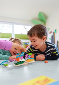 Kleurige, in elkaar grijpende schuimvormen in verschillende felle kleuren, gestapeld op een witte ondergrond, met kinderen die spelen aan een tafel.