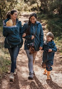 Zwei Frauen gehen auf einem Schotterweg und tragen blaue Jacken. Eine von ihnen trägt ein Baby in einer Tasche; ein Junge mit gelben Stiefeln hält ein Fernglas. Natürliche Umgebung.