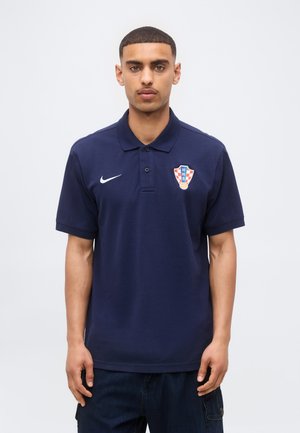 Jeune homme aux cheveux courts portant un polo bleu marine avec le logo Nike et l'écusson du football croate, debout sur un fond blanc uni.
