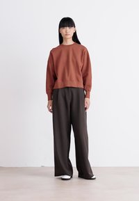 Mujer de pie con una blusa de manga larga color óxido, pantalones anchos sueltos de un marrón oscuro y zapatos blancos, frente a un fondo claro y liso.