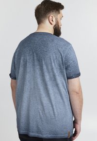 Blå gradient t-shirt med rund halsudskæring, korte rullede ærmer og en tekstureret overflade. Bagsiden viser subtile falmninger og et side-label.