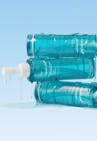 Huile démaquillante dans des bouteilles en plastique transparent bleu sarcelle avec pompes blanches. L'étiquette comprend le nom du produit et les instructions. Du liquide s'écoule des bouteilles.