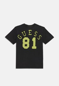 Black t-shirt featuring yellow-green raised text "GUESS 81" on the back. Cotton material, short sleeves, round neckline, casual fit.