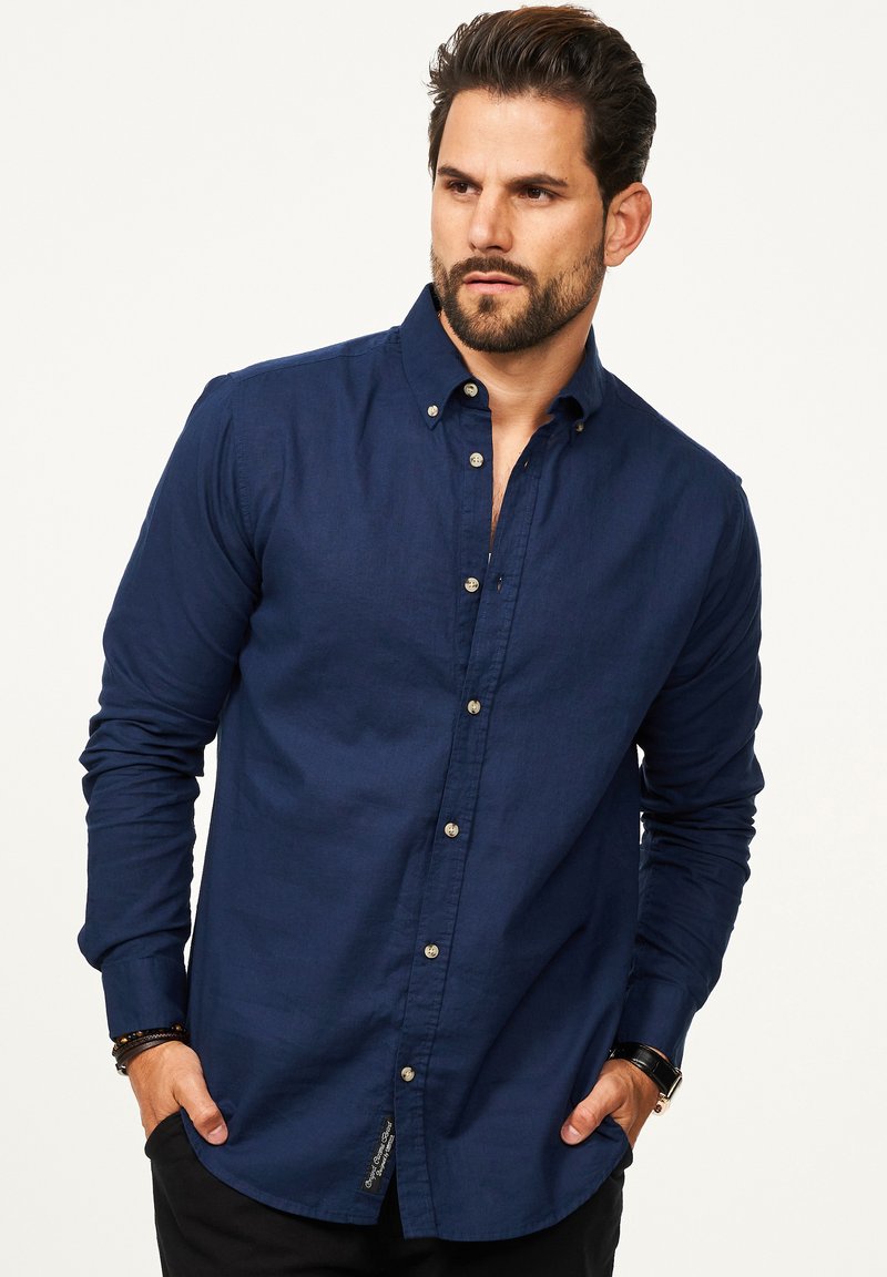 Marineblaue langärmlige Knopfleiste mit Kragen, mit kontrastierenden Knöpfen und einem Etikett unten links. Glatte Stofftextur.