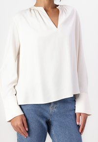 Blouse à manches longues couleur crème avec un col en V et un col froncé, conçue dans un tissu fluide et léger. Associée à un jean en denim bleu.