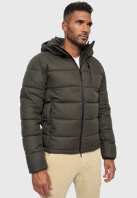 Grüne gepolsterte Jacke mit hohem Kragen und Kapuze, ausgestattet mit einem Reißverschluss und zwei seitlichen Taschen, gestaltet aus strukturiertem Stoff.