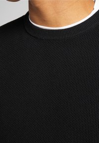 Schwarzer gerippter Strickpullover, der eine Nahaufnahme des strukturierten Stoffs und des Rundhalsausschnitts zeigt, der über einem weißen Kragen liegt.