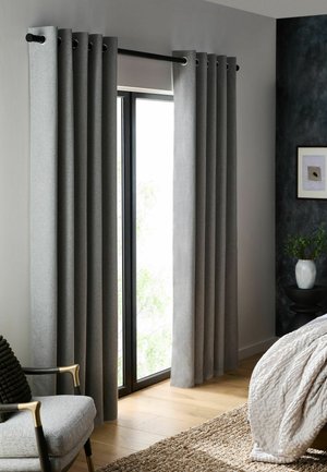 Light gray curtains hang from a black rod over a large glass door in a bedroom with wooden floor and light bedding.