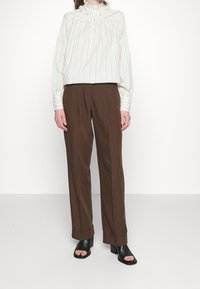 Vit och ljusgrönrandig blus med lång ärm och volangkrage, tillsammans med bruna veckade byxor och svarta klacksandaler.