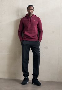 Homme appuyé contre un mur beige, portant un sweat à capuche maroon de Puma, un pantalon de survêtement noir de Puma avec texte logo, et des baskets noires de Puma.