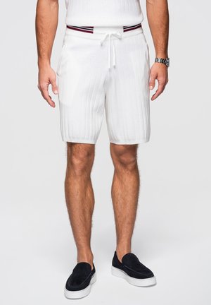 Mand iført hvide ribstrikkede shorts med snøre og stribet linning, sorte slip-on sko og sølvfarvet armbåndsur på venstre håndled.