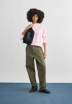 Kvinde med krøllet hår iført rosa sweatshirt og olivengrønne bukser, med en sort skuldertaske over den ene skulder, stående på blå tæppe.