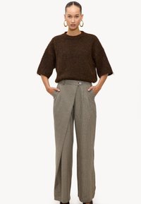 Mujer con un suéter corto de manga corta de color marrón oscuro y peludo, con pantalones de talle alto de pierna ancha a cuadros en beige y marrón, con las manos en los bolsillos.