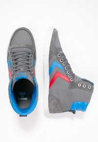 Grå højtop sneakers med blå og røde accenter. Funktioner inkluderer en tekstureret overdel, polstret krave, hvid gummisål og kontrasterende chevron-design.