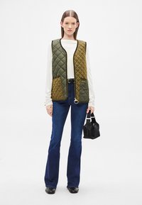 Πλεκτό γιλέκο σε ελιά και μουσταρδί, με φερμουάρ στο μπροστινό μέρος, δύο τσέπες και στρογγυλό τσεκ για το φερμουάρ, σε συνδυασμό με τζιν καμπάνα και μαύρη τσάντα.