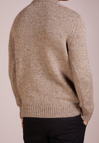 Pull en tricot de couleur beige clair, avec un motif texturé, des poignets et un ourlet côtelés, et une coupe décontractée. Porté par-dessus un pantalon foncé.