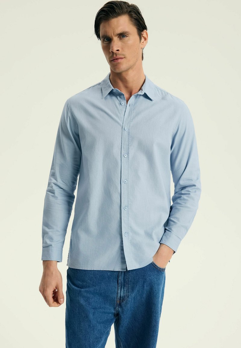 Jasnoniebieska koszula z długim rękawem, wykonana z teksturowanej tkaniny, z kołnierzykiem zapinanym na guziki oraz guzikami z przodu, w połączeniu z niebieskimi dżinsami.