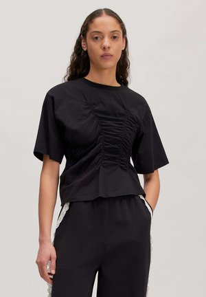 Vrouw met lang krullend haar die een zwarte gerimpelde top met korte mouwen en een zwarte broek met witte kantafwerking op de zakken draagt.