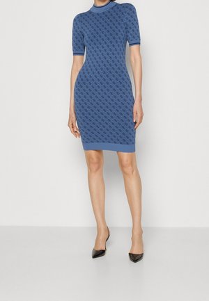 Femme portant une robe ajustée bleu à motifs longueur genou avec des manches courtes et des talons noirs à bout pointu, debout contre un fond uni.