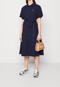 Mörkblå kortärmad skjortklänning med knäppning och midjeband. Har en liten logotyp på bröstet och en sluttande kjol. Vita sneakers och en beige handväska.