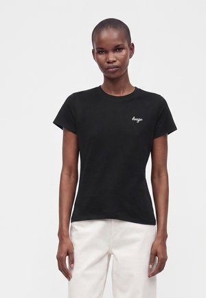 Femme aux cheveux courts portant un t-shirt noir ajusté avec un texte blanc "hugo" et un pantalon blanc, debout devant un fond clair uni.