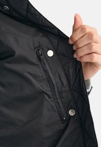 Black quilted jacket interior with a visible zip pocket and two snap buttons. The fabric is smooth, lightweight, and matte in texture.