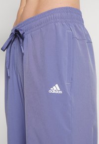 Pantalon de sport violet en tissu léger, doté d'une taille élastique avec cordon de serrage et d'un logo Adidas blanc sur la cuisse.